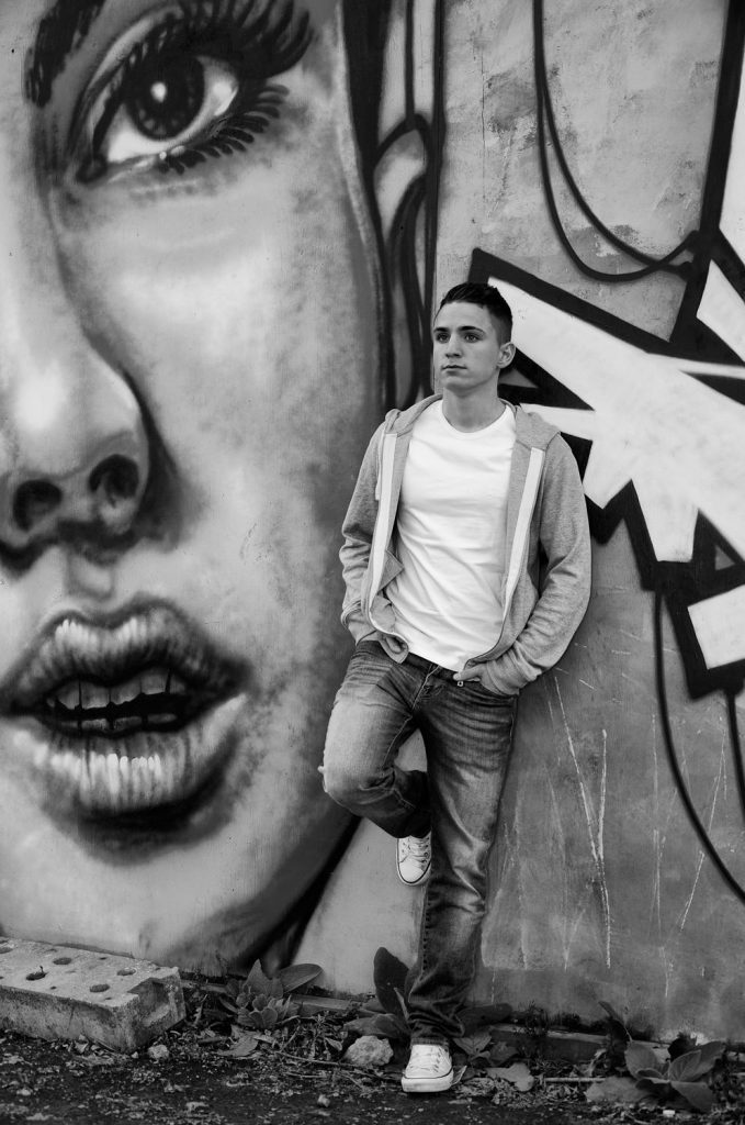 Schwarz-Weiß-Foto: junger Mann vor einer Graffiti-Wand mit großem Gesicht